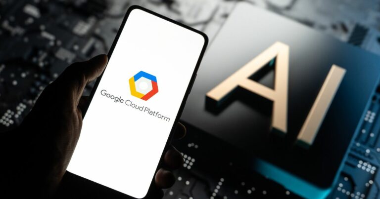 Uma mão segura um smartphone que exibe o logotipo e o texto da Google Cloud Platform. Ao fundo, vê-se a palavra "AI" (inteligência artificial) em destaque sobre uma superfície que lembra um circuito eletrônico ou uma placa tecnológica, sugerindo um ambiente digital e de alta tecnologia.