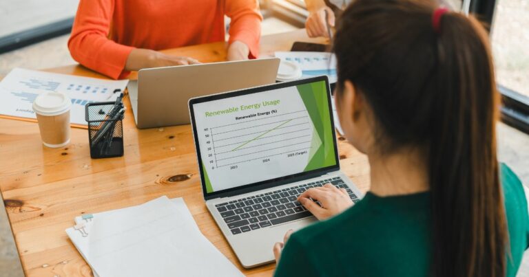 Três pessoas estão sentadas em uma mesa de madeira, trabalhando em laptops em busca de oportunidades. Uma delas veste uma camisa laranja, outra uma camisa verde, e a terceira pessoa está parcialmente visível. O laptop da pessoa com camisa verde exibe um gráfico intitulado "Uso de Energia Renovável", com dados de 2010 a 2020. Sobre a mesa, há também alguns papéis e uma xícara de café.
