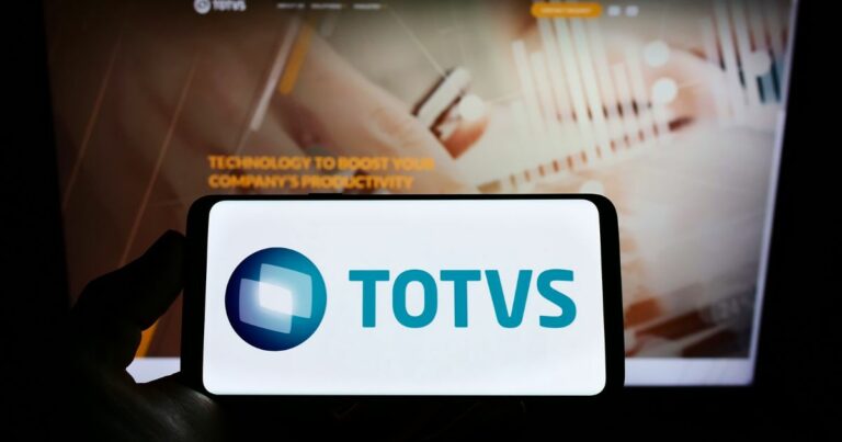 Um smartphone exibindo o logotipo da TOTVS, composto por um design geométrico azul e branco ao lado do texto 'TOTVS' em azul. Ao fundo, há uma tela de computador desfocada com o texto 'TECHNOLOGY TO BOOST YOUR COMPANY'S PRODUCTIVITY.