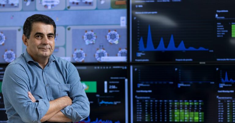 Reinaldo Sima, executivo da MRV, posa com os braços cruzados diante de telas com gráficos e painéis de dados, em ambiente corporativo de monitoramento tecnológico.