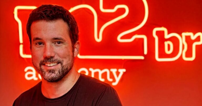 Imagem de Ricardo Marsili, especialista em inteligência artificial, sorrindo em frente a uma parede vermelha iluminada com letreiro em neon da M2BR Academy. Ele veste uma camiseta preta e está em um ambiente com iluminação quente, destacando o logotipo ao fundo. (Ricardo Marsili, M2BR, Brasil)