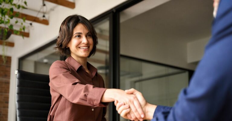 Mulher sorridente com cabelo castanho escuro e curto, vestindo uma camisa marrom, está sentada em uma cadeira de escritório e cumprimentando alguém com um aperto de mão. A cena parece ocorrer em um ambiente corporativo moderno, com paredes de vidro e vigas de madeira no teto. A pessoa com quem ela aperta a mão veste um terno azul, mas está fora de foco e parcialmente fora do enquadramento. A atmosfera transmite cordialidade e profissionalismo, sugerindo uma entrevista de emprego ou reunião de negócios. (vagas)