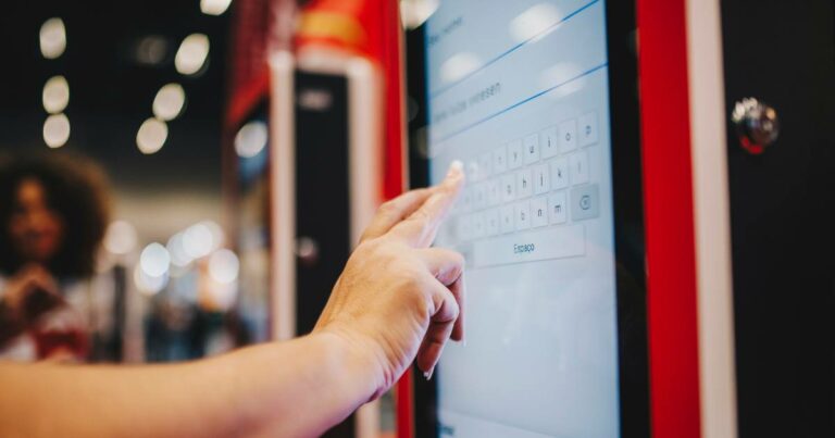 Pessoa interage com um totem eletrônico do iFood, com a interface da empresa Videosoft visível na tela. A mão está digitando em um teclado virtual exibido no display sensível ao toque. O ambiente ao fundo está desfocado, sugerindo um local público movimentado, como um shopping ou praça de alimentação