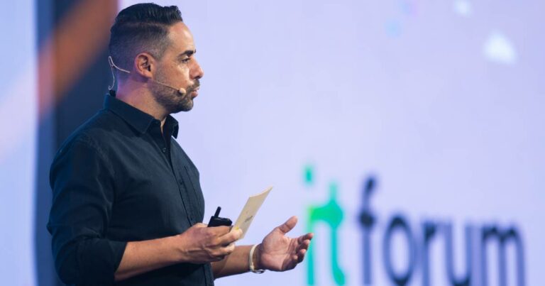André Cavalli, CEO do IT Forum, aparece de perfil durante uma apresentação no palco, segurando um microfone de mão e um papel, com microfone auricular acoplado. Ao fundo, está o logotipo do evento "IT Forum" projetado em uma tela.