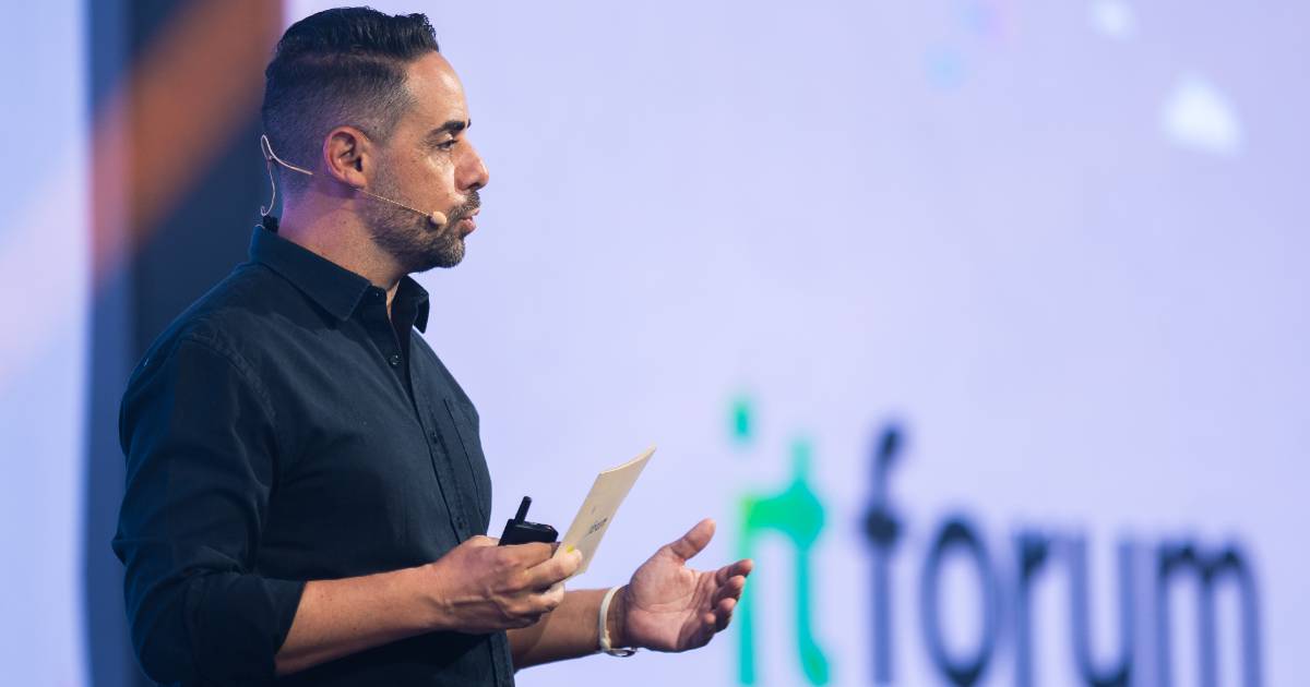 André Cavalli, CEO do IT Forum, aparece de perfil durante uma apresentação no palco, segurando um microfone de mão e um papel, com microfone auricular acoplado. Ao fundo, está o logotipo do evento "IT Forum" projetado em uma tela.