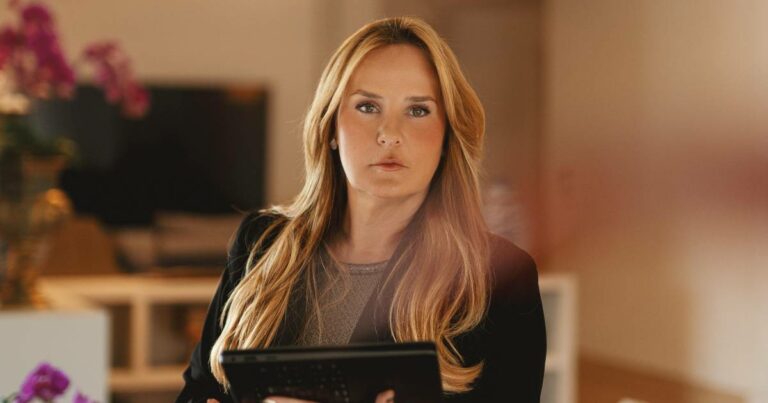 Mulher loira de cabelos longos e lisos, vestindo roupa social escura, segura um tablet e olha diretamente para a câmera. Ao fundo, há um ambiente interno com móveis e flores decorativas. Na imagem está Flávia Pollo Nassif, CEO da DMA