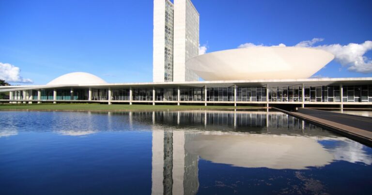 A imagem mostra o Congresso Nacional do Brasil, em Brasília. O complexo arquitetônico é composto por duas torres altas ladeadas por duas cúpulas — uma em cada lado. A estrutura está refletida em um espelho d’água no primeiro plano da imagem. (governo)