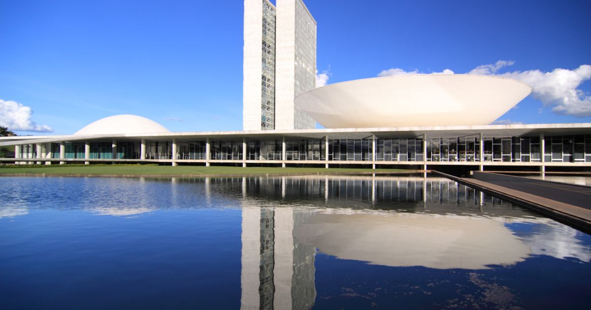 A imagem mostra o Congresso Nacional do Brasil, em Brasília. O complexo arquitetônico é composto por duas torres altas ladeadas por duas cúpulas — uma em cada lado. A estrutura está refletida em um espelho d’água no primeiro plano da imagem. (governo)