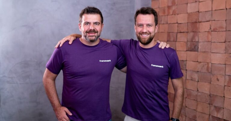 Duas pessoas estão em pé, lado a lado, com os braços apoiados nos ombros uma da outra. Ambas vestem camisetas roxas com a palavra "framework" estampada na frente. Ao fundo, há uma parede cinza à esquerda e uma parede de tijolos à direita.