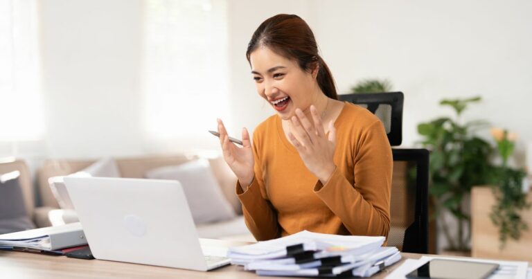 Uma pessoa está sentada à mesa com um laptop aberto, várias pilhas de papéis e um tablet. Ao fundo, há um sofá e algumas plantas decorativas. Não há problemas de matemática ou tarefas escolares visíveis na imagem. (vagas, emprego)
