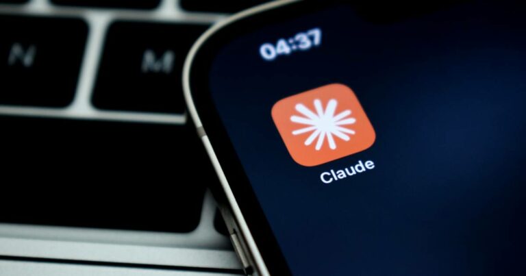 Ícone do aplicativo Claude, da empresa Anthropic, exibido na tela de um smartphone. O ícone é laranja com um símbolo branco estilizado no centro, lembrando um asterisco ou explosão, com o nome "Claude" escrito abaixo. Ao fundo, vê-se parcialmente o teclado de um laptop. IBM