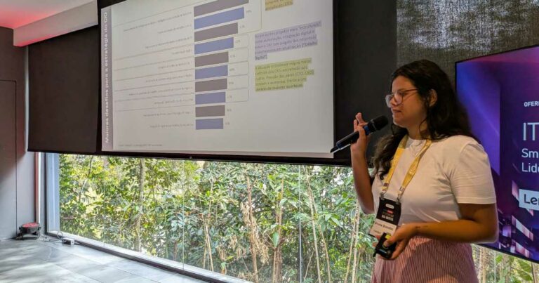 Bruna Bomfim, gerente de Inteligência da Itaqui, apresentando palestra durante o IT Forum Na Mata, em frente a um telão com gráficos e segurando um microfone. (investimentos)