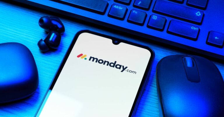 Um smartphone está sobre uma mesa, com a tela exibindo o logotipo e o nome "monday.com" em texto preto, acompanhado por três pontos coloridos (vermelho, amarelo e azul) dispostos diagonalmente. Ao redor do celular, há um teclado de computador, um mouse sem fio e fones de ouvido também sem fio. A cena é iluminada com luz azul, sugerindo um ambiente de trabalho moderno e tecnológico.