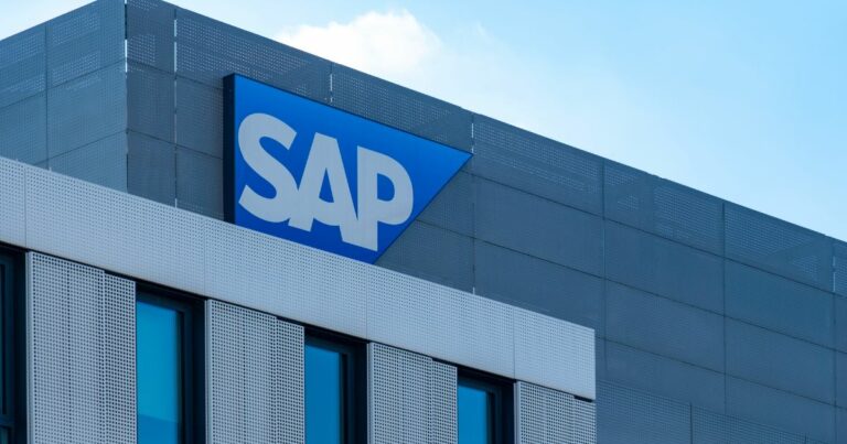 Essa imagem mostra a fachada de um prédio moderno com o logotipo da SAP em destaque na parte superior — letras brancas sobre um fundo azul em formato triangular. A construção tem janelas amplas e painéis metálicos perfurados, conferindo um visual contemporâneo e sofisticado. Ela parece representar uma sede ou escritório da SAP, empresa multinacional conhecida por desenvolver softwares corporativos para gestão de operações e relacionamento com clientes.