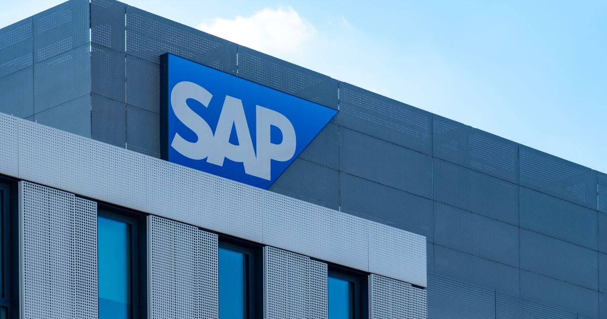 Essa imagem mostra a fachada de um prédio moderno com o logotipo da SAP em destaque na parte superior — letras brancas sobre um fundo azul em formato triangular. A construção tem janelas amplas e painéis metálicos perfurados, conferindo um visual contemporâneo e sofisticado. Ela parece representar uma sede ou escritório da SAP, empresa multinacional conhecida por desenvolver softwares corporativos para gestão de operações e relacionamento com clientes.