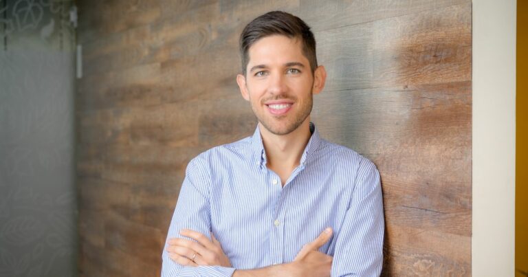 Uma pessoa está em pé com os braços cruzados, posicionada diante de uma parede de madeira. Ela veste uma camisa azul clara com listras e tem cabelo curto. À esquerda, há uma seção de vidro fosco com padrões visíveis. A imagem transmite um ambiente casual e profissional, ideal para uso em perfis institucionais ou materiais promocionais. (Nuvemshop)