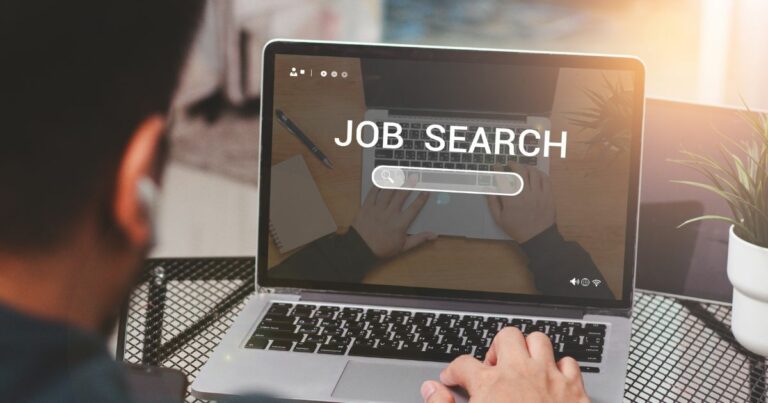 Pessoa utilizando um laptop, com as mãos posicionadas sobre o teclado. Na tela do computador aparece uma barra de pesquisa com o texto "JOB SEARCH" acima dela, indicando uma busca por emprego. Ao lado do laptop, há um caderno aberto e uma caneta, sugerindo que a pessoa está tomando notas ou se organizando durante a pesquisa. (vagas)