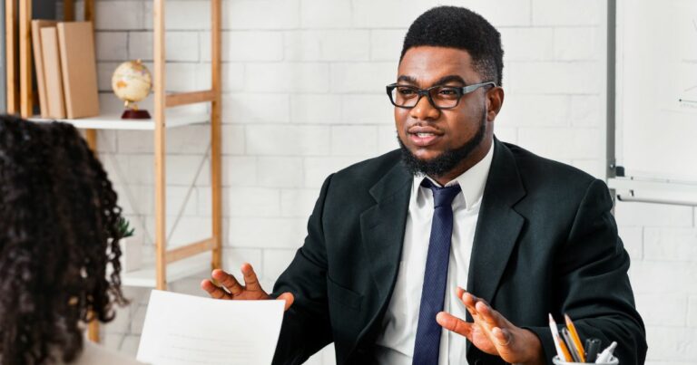 Um homem vestido com terno e gravata, usando óculos e com cabelo e barba bem aparados, está em um ambiente profissional, aparentemente envolvido em uma conversa com outra pessoa que está de costas para a câmera. Ao fundo, há prateleiras com livros, um globo terrestre e parte de uma lousa branca visível à direita. A cena sugere uma reunião de negócios ou uma entrevista em um escritório bem organizado. (emprego/vagas)