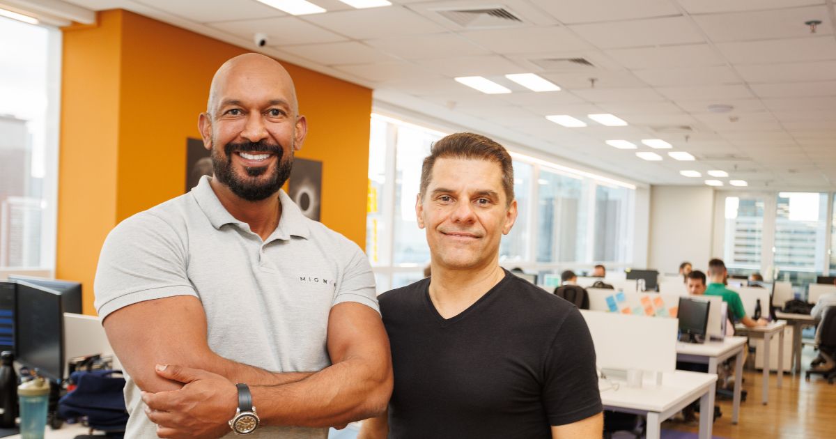 Duas pessoas estão em pé, lado a lado, em um escritório moderno e bem iluminado. A pessoa à esquerda veste uma camisa polo cinza claro com o logotipo "MIGN" e está com os braços cruzados. A pessoa à direita usa uma camiseta preta. Ao fundo, há várias mesas com computadores e outras pessoas trabalhando. Grandes janelas permitem a entrada de luz natural e revelam uma vista urbana. O ambiente transmite a ideia de um espaço colaborativo ou escritório corporativo. (Mignow)