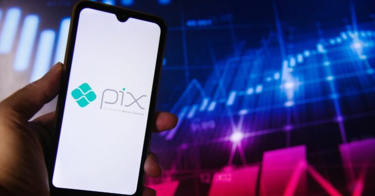 Uma mão segura um smartphone cuja tela exibe o logotipo do "Pix", acompanhado do texto "powered by Banco Central". Ao fundo, há um gráfico digital com linhas e barras, sugerindo dados financeiros ou informações do mercado de ações.