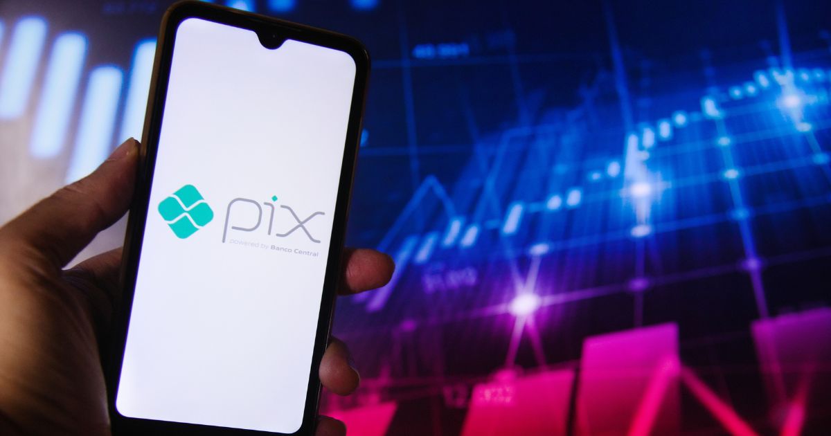 Uma mão segura um smartphone cuja tela exibe o logotipo do "Pix", acompanhado do texto "powered by Banco Central". Ao fundo, há um gráfico digital com linhas e barras, sugerindo dados financeiros ou informações do mercado de ações.