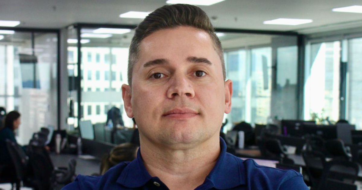 Pessoa com cabelo curto, vestindo uma camisa polo azul escura, em um ambiente de escritório. Ao fundo, há móveis de escritório, janelas com vista para a cidade e outras pessoas trabalhando em suas mesas. (Opah IT)