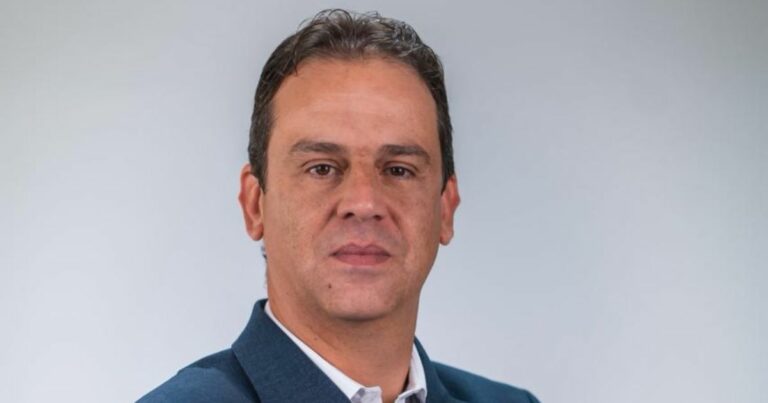 Pessoa em ambiente formal, vestindo paletó azul escuro sobre camisa branca. O fundo é claro e simples, destacando a figura principal. A imagem pode ser usada em contextos profissionais ou institucionais, como identificação ou apresentação. (RH)