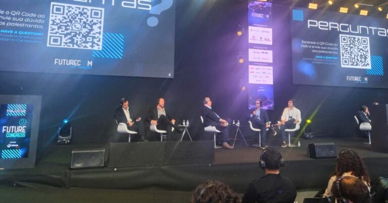 A imagem mostra um painel de discussão durante o evento Future Congress. Cinco pessoas estão sentadas no palco, cada uma com um microfone, participando da conversa. Ao fundo, duas telas grandes exibem a palavra "PERGUNTAS?", acompanhada de um QR code e instruções para que o público envie perguntas aos palestrantes. O cenário inclui logotipos e elementos visuais relacionados à identidade do evento. Na parte frontal da imagem, é possível ver o público assistindo atentamente. (logistica)