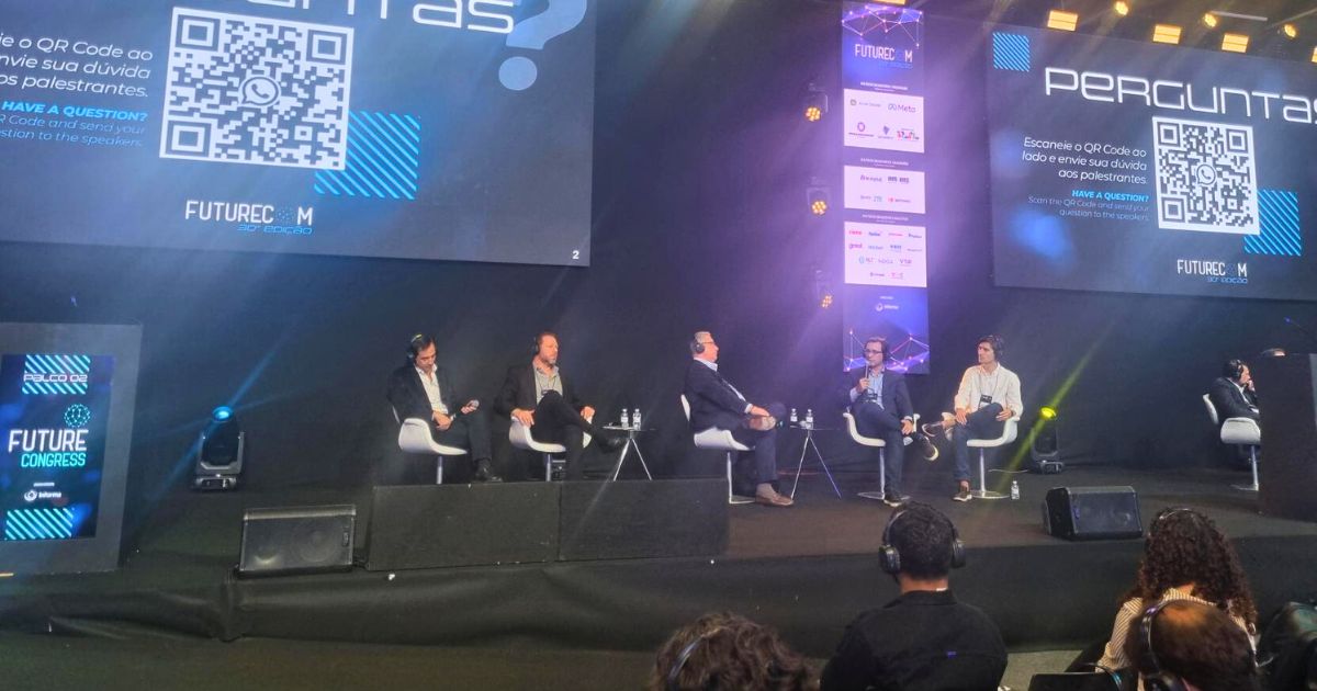 A imagem mostra um painel de discussão durante o evento Future Congress. Cinco pessoas estão sentadas no palco, cada uma com um microfone, participando da conversa. Ao fundo, duas telas grandes exibem a palavra "PERGUNTAS?", acompanhada de um QR code e instruções para que o público envie perguntas aos palestrantes. O cenário inclui logotipos e elementos visuais relacionados à identidade do evento. Na parte frontal da imagem, é possível ver o público assistindo atentamente. (logistica)