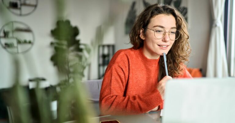 Uma pessoa com cabelo cacheado e óculos está sentada à mesa, vestindo um suéter laranja. Ela segura uma caneta próxima à boca enquanto observa atentamente a tela de um laptop à sua frente. Ao fundo, é possível ver um ambiente interno desfocado, com plantas e prateleiras na parede, sugerindo um espaço de trabalho ou estudo. A imagem transmite concentração e envolvimento, possivelmente durante uma reunião online ou momento de estudo de cursos.