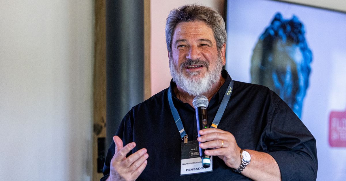 A imagem mostra uma pessoa com cabelo grisalho e barba, vestindo uma camisa preta e segurando um microfone. Ela está falando ou apresentando, com gestos visíveis. A pessoa usa um crachá com as palavras “PENSADOR” e “MARCO ANTONIO VILLA”. Ao fundo, há uma tela desfocada exibindo o que parece ser uma escultura ou obra de arte. (tecnologia)