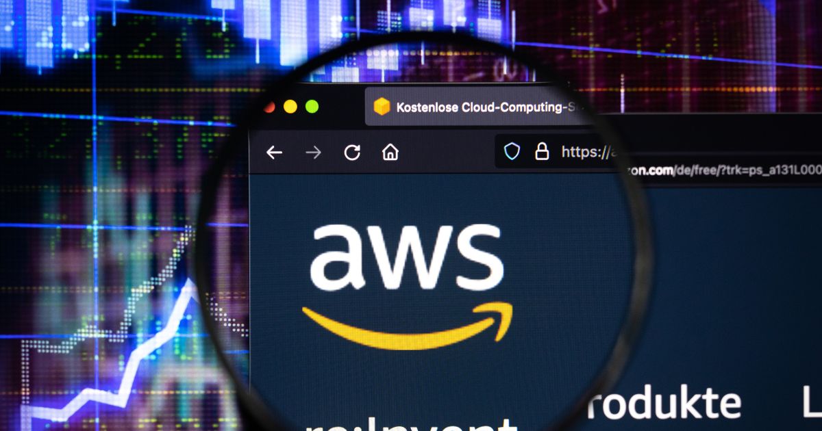 A imagem mostra uma tela de computador exibindo o site da Amazon Web Services (AWS). O logotipo da AWS, com as letras em branco e uma seta curva amarela abaixo, está ampliado por uma lupa no centro da imagem. Na parte superior, é visível a barra do navegador com um título em alemão: “Kostenlose Cloud-Computing-S…”. Ao fundo, aparecem gráficos digitais em tons de azul e roxo, sugerindo um contexto tecnológico ou financeiro.