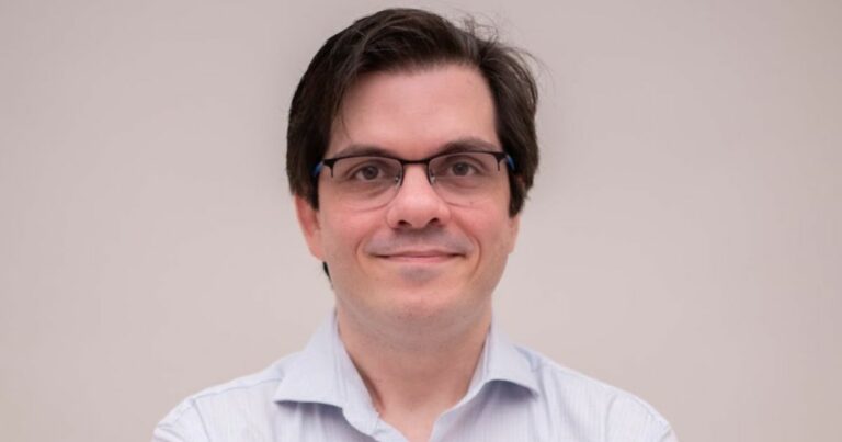 A imagem mostra uma pessoa vestindo uma camisa social de cor clara, com colarinho e botões visíveis. O cabelo é escuro e está penteado para trás, com um aspecto levemente volumoso. O fundo é simples e uniforme, em tom claro, sugerindo um ambiente neutro ou estúdio fotográfico. A pessoa está posicionada de frente, com os braços cruzados à altura do peito. ( Flutter Brazil )