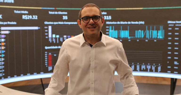 Pessoa em pé diante de uma grande tela que exibe diversos gráficos e visualizações de dados. A tela apresenta gráficos de barras e de linhas com títulos como “Ticket Médio”, “Total de Clientes”, “Clientes que Mais Compraram”, “Histórico de Diário de Vendas (VV)” e “Últimas Vendas”. Alguns valores visíveis incluem R$29,32 como ticket médio, 35 como total de clientes e 315 como outro indicador não especificado. A imagem retrata um ambiente profissional voltado à apresentação de métricas de desempenho, possivelmente relacionadas a vendas ou análise de negócios. (inteligência artificial, IA, empresas)