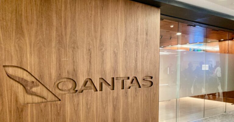 A imagem mostra uma parede de madeira com o logotipo da Qantas gravado, incluindo um canguru estilizado — símbolo da companhia aérea australiana. O nome “Qantas” também está gravado ao lado do logotipo. Ao fundo, há portas de vidro e reflexos de pessoas, sugerindo que o local pode ser um escritório ou uma sala de espera.