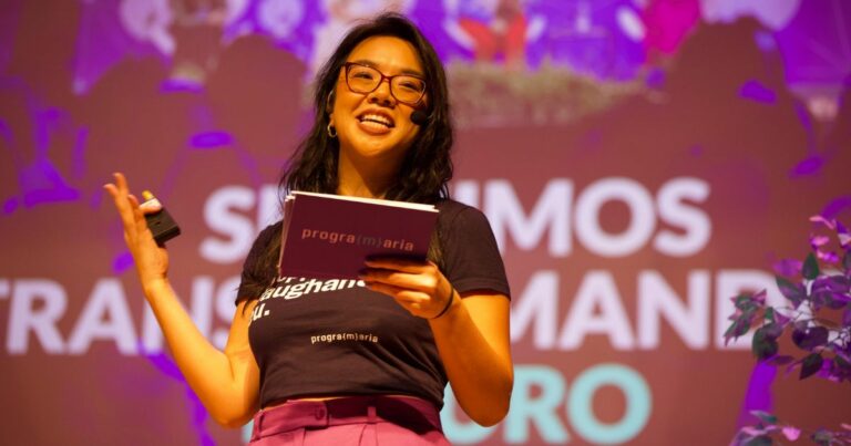 A imagem mostra uma pessoa em pé, segurando um microfone ou controle em uma mão e um caderno ou bloco de anotações na outra. A pessoa veste uma camiseta preta com o texto visível “programaria” e outras palavras parcialmente legíveis. Ao fundo, há um painel iluminado com letras grandes, onde se lê “SOMOS TRANS...” e outras palavras desfocadas, sugerindo um evento ou palestra. A iluminação é intensa, com tons roxos e rosados predominando, e há elementos decorativos como plantas no canto inferior direito. (diversidade)