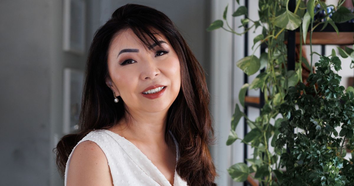 A imagem mostra uma pessoa com cabelo escuro e liso, caindo sobre os ombros. Está vestindo uma blusa branca sem mangas, com textura leve e discreta. Ao fundo, há uma parede clara e uma estante com plantas verdes, criando um ambiente interno com decoração natural e iluminação suave. (tecnologia)