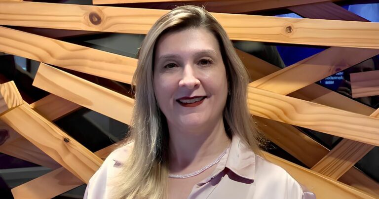 A imagem mostra uma pessoa vestindo uma camisa de tecido acetinado em tom claro, com gola tradicional e botões parcialmente visíveis. Ao redor do pescoço, há um colar delicado, composto por pequenas contas ou pedras claras. O fundo é formado por uma estrutura artística de ripas de madeira dispostas em ângulos cruzados, criando um padrão geométrico interessante. Entre as ripas, é possível ver áreas escuras e reflexos que sugerem um ambiente interno com iluminação controlada. (transforma)