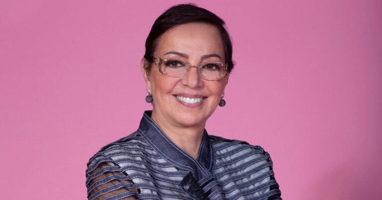 A imagem mostra uma pessoa vestindo uma jaqueta estilosa de tecido semelhante ao jeans, com um design moderno composto por faixas horizontais translúcidas nas mangas e no corpo. A peça tem gola alta estruturada e detalhes que criam um visual sofisticado e contemporâneo. O fundo é totalmente rosa, liso e uniforme, destacando ainda mais a roupa e criando um contraste vibrante. A pessoa está posicionada de frente, com os braços cruzados, transmitindo uma postura firme e elegante. (Tânia)