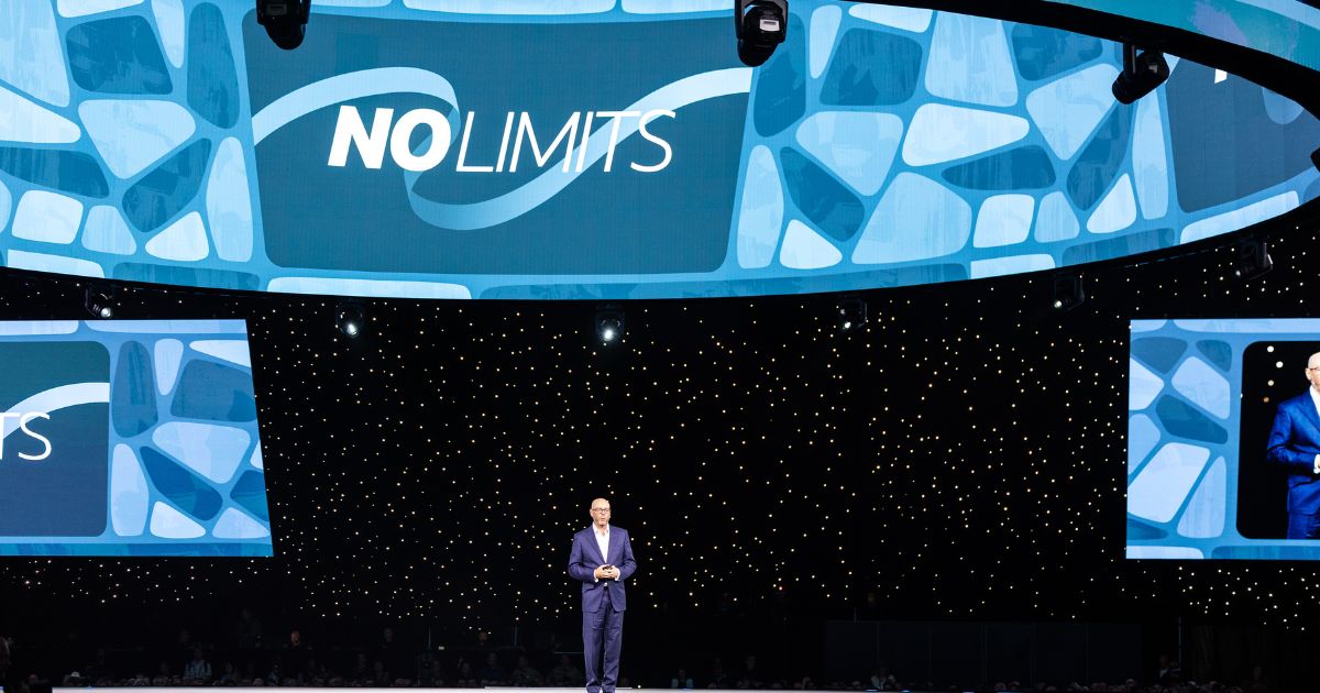 Pessoa em pé sobre um palco, com um grande telão ao fundo exibindo a frase “NO LIMITS” sobre um fundo abstrato em tons de azul. Há também duas telas menores nas laterais, mostrando a mesma mensagem e padrão visual. O cenário é complementado por diversas luzes pequenas ao fundo, lembrando um céu estrelado. A imagem sugere um evento ou apresentação com a temática de superação ou possibilidades ilimitadas. (NetSuite)