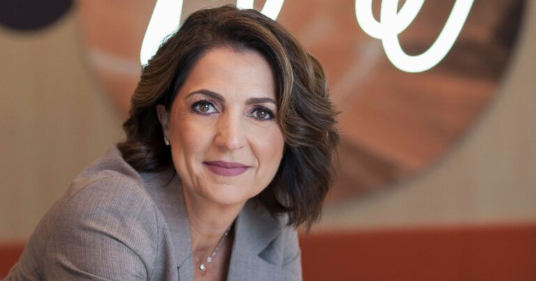 Pessoa com cabelo curto e ondulado, vestindo blazer cinza, olha diretamente para a câmera. Ao fundo, há um objeto circular desfocado com texto cursivo branco parcialmente legível. A imagem apresenta um estilo de retrato profissional, com foco na pessoa em primeiro plano. (AWS)