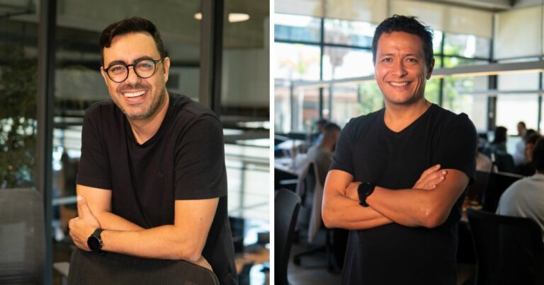 A imagem mostra duas pessoas lado a lado em um ambiente de escritório moderno. Ambas estão usando camisetas pretas e óculos, e posam sorrindo com os braços cruzados. Ao fundo, é possível ver paredes de vidro, mesas e outras pessoas trabalhando, sugerindo um espaço corporativo colaborativo. (fintech)