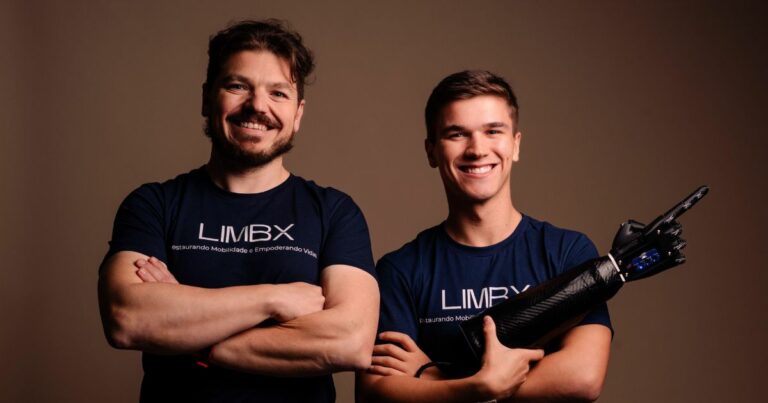 Duas pessoas estão em pé lado a lado, vestindo camisetas azul-escuras com a palavra "LIMBX" estampada na frente. A pessoa à esquerda está com os braços cruzados, enquanto a pessoa à direita segura uma prótese de braço. O fundo é simples, em tom marrom. A imagem destaca tecnologia avançada em próteses, possivelmente relacionada a uma iniciativa de inclusão ou mobilidade.