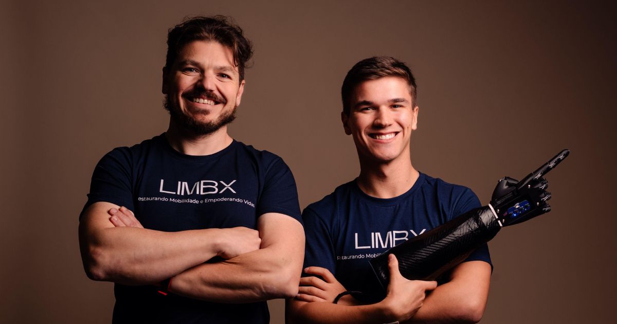 Duas pessoas estão em pé lado a lado, vestindo camisetas azul-escuras com a palavra "LIMBX" estampada na frente. A pessoa à esquerda está com os braços cruzados, enquanto a pessoa à direita segura uma prótese de braço. O fundo é simples, em tom marrom. A imagem destaca tecnologia avançada em próteses, possivelmente relacionada a uma iniciativa de inclusão ou mobilidade.