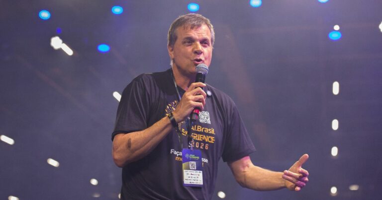A cena mostra uma pessoa em pé sobre um palco, segurando um microfone na mão direita e gesticulando com a mão esquerda. A pessoa veste uma camiseta preta com texto em amarelo e branco que inclui as palavras “AI Brasil Experience 2026” e um crachá pendurado no pescoço com QR code e logotipos de patrocinadores. O fundo está iluminado com luzes roxas e azuis, criando um efeito de evento tecnológico ou conferência, com vários pontos de luz desfocados ao fundo, sugerindo um ambiente amplo e moderno. (IA)