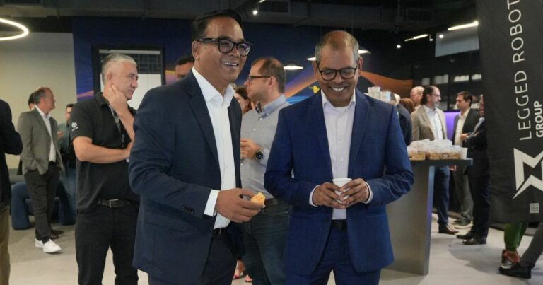 A imagem mostra um grupo de pessoas em um ambiente profissional, possivelmente durante um evento de networking ou conferência. No primeiro plano, duas pessoas vestindo ternos estão conversando e segurando bebidas. Ao fundo, outros participantes também interagem e consomem petiscos. À direita, há um banner com o texto "LEGGED ROBOT GROUP", sugerindo que o evento esteja relacionado à área de robótica ou tecnologia. O ambiente é interno, com iluminação moderna e decoração contemporânea. (TCS)