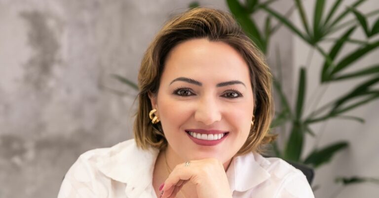 A imagem mostra uma pessoa com cabelo curto, vestindo uma camisa branca e brincos dourados. Ao fundo, há uma parede desfocada e plantas verdes, sugerindo um ambiente interno com elementos naturais. A composição destaca a pessoa em um contexto profissional e bem iluminado. (startups e escalabilidade)