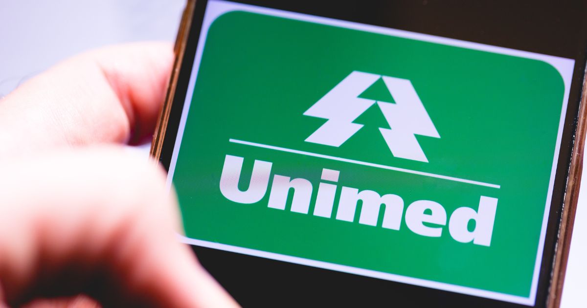 A imagem mostra um close de uma tela de smartphone exibindo o logotipo da Unimed, uma cooperativa médica e operadora de plano de saúde brasileira. O logotipo tem fundo verde, com um símbolo branco que lembra duas árvores ou setas apontando para cima, e a palavra “Unimed” escrita em letras brancas em destaque abaixo do símbolo, WhatsApp