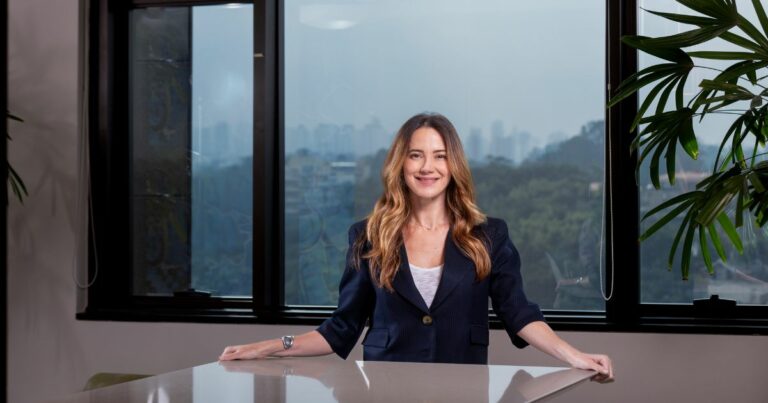 A imagem mostra uma pessoa em pé atrás de uma mesa, em um ambiente de escritório. Ela veste um blazer escuro sobre uma blusa clara e tem cabelo longo e ondulado. Ao fundo, há grandes janelas que revelam uma paisagem urbana com áreas verdes, além de plantas internas visíveis à direita. A composição transmite um contexto profissional, ideal para ilustrar temas relacionados a negócios ou ambientes corporativos. (Gisselle, liderança)