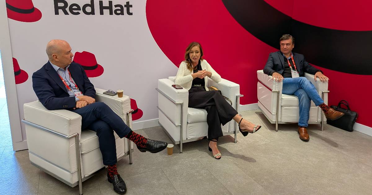 Três executivos da Red Hat estão sentados em poltronas brancas, participando de uma conversa em um ambiente corporativo com o logotipo da Red Hat ao fundo. Da esquerda para a direita: Gilson Magalhães, vice-presidente e gerente geral da Red Hat para a América Latina; Sandra Vaz, presidente da Red Hat Brasil; e Alexandre Duarte, vice-presidente de Serviços.